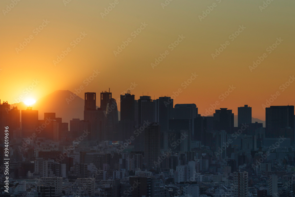 Naklejka premium 夕陽が沈むオレンジの空を背景にシルエットになって見える富士山と新宿のビル群