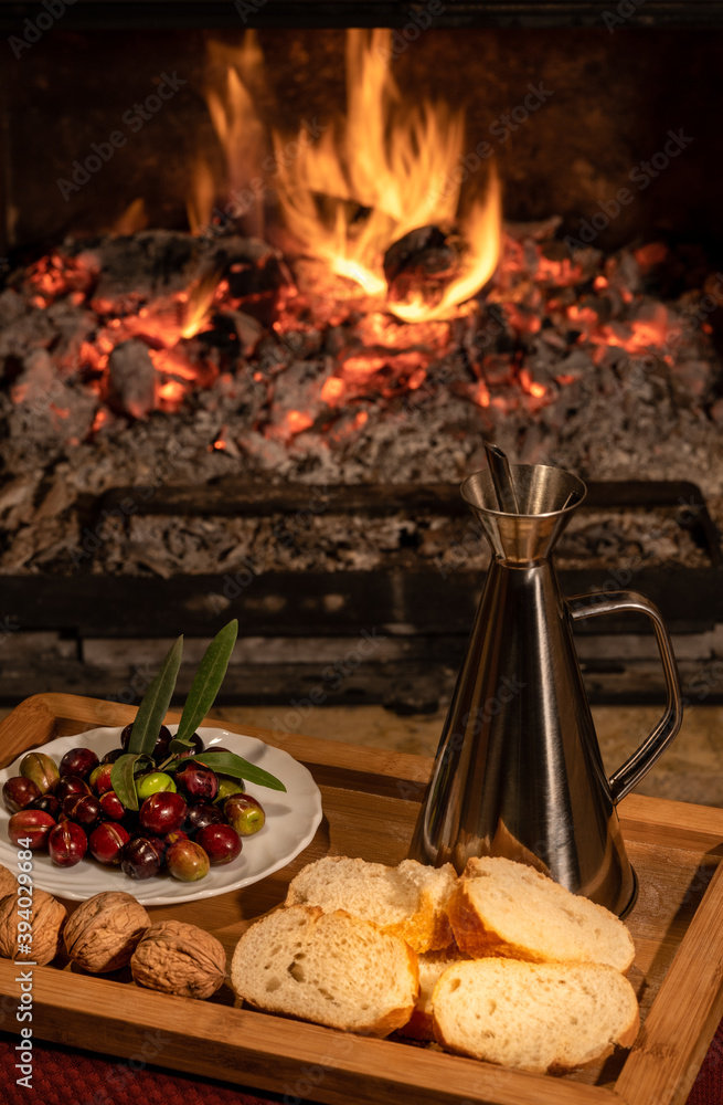 Aluminium oil cruet with flavoured olive oil on a wooden tray accompanied by raw olives, bread and nuts, over a rustic fire