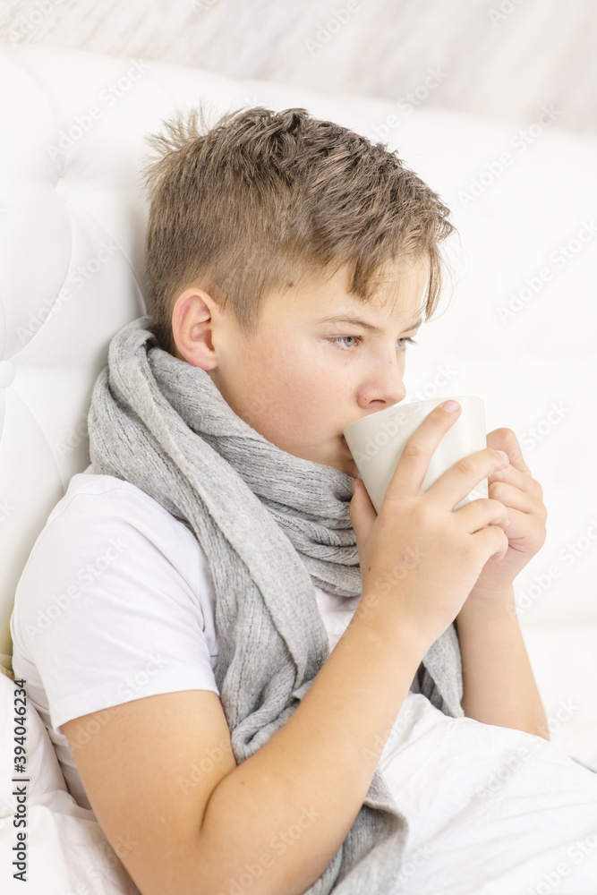 A sick child lies on a bed in a warm scarf and drinks hot tea from a mug in the beadroom. An unhealthy child drinks from mug. Cold season.