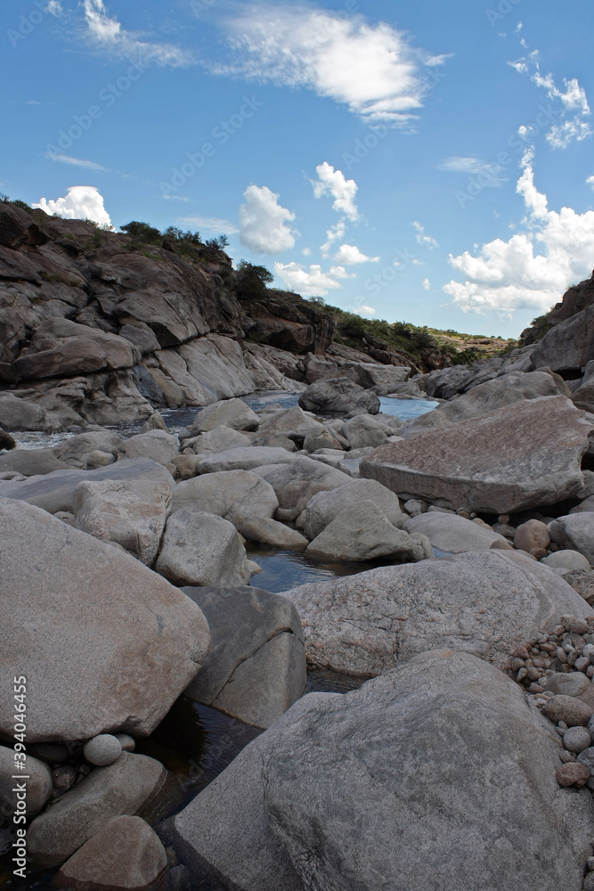 View of the rocky river flowing across the stone valley in the mountain forest. The shallows, rocky shore and bed under a blue sky in a summer sunny day. View of the rocky river flowing across the stone valley in the mountain forest. The shallows, rocky shore and bed under a blue sky in a summer sunny day.