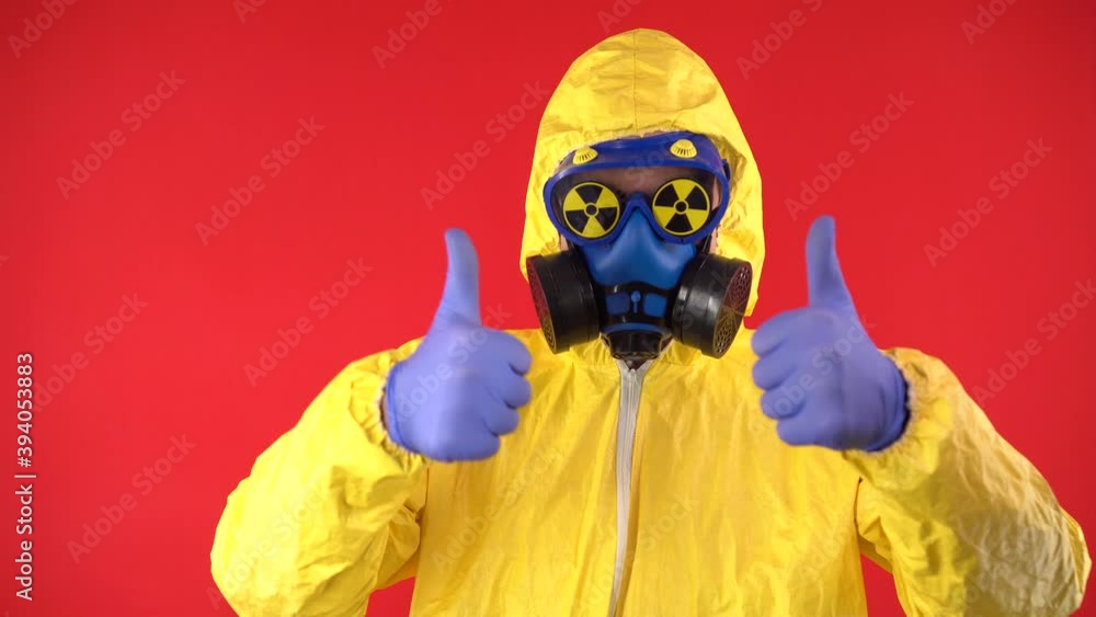 A man in a protective chemical suit and mask with signs of radiation in glasses shows a thumbs up. Isolate, studio shooting on a red background. The concept of radiation and hazard. A man in a protective chemical suit and mask with signs of radiation in glasses shows a thumbs up. Isolate, studio shooting on a red background. The concept of radiation and hazard.