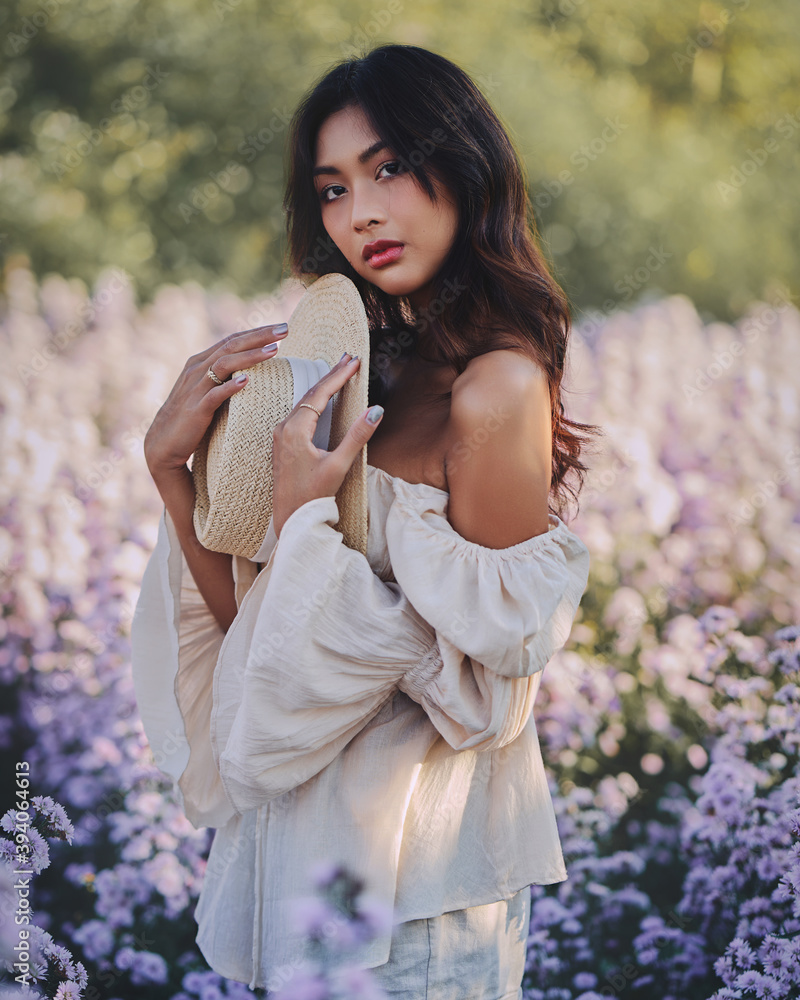 Lifestyle fashion portrait of stylish young asian woman with straw hat posing at the flowers field . Wearing straw hat and beige outfit .Summer bright colors.