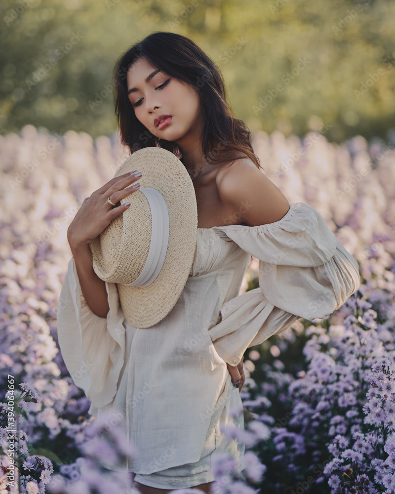 Lifestyle fashion portrait of stylish young asian woman with straw hat posing at the flowers field . Wearing straw hat and beige outfit .Summer bright colors.