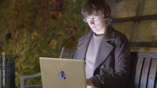 Hipster sitting in the park at night and typing on laptop. Young man working outdoors. Autumn evening. Distant work. Steadicam shot