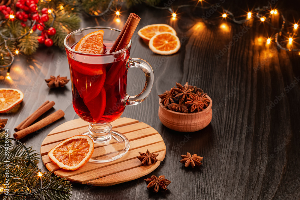 Mulled wine in a beautiful glass with orange and cinnamon on a dark background with fir branches, garland and red berries. Nearby are orange slices, cinnamon, anise and spruce branches. Copy space.
