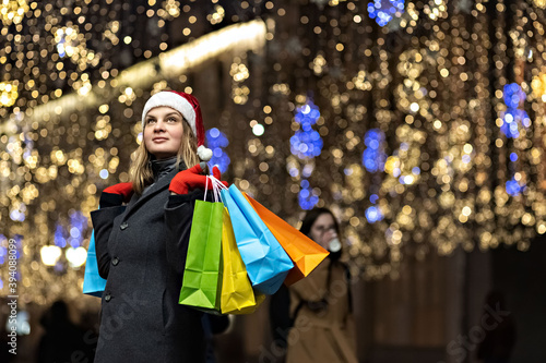 A woman with long hair and a Santa hat near the window of a city store with purchases in colorful, paper bags. New year's shopping