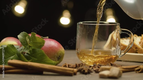 Close-up hot tea pouring into glass mug on black background. Steam rises above the cup. Fresh apples with leaves and spices on the table. Slide or move camera. Dolly shot