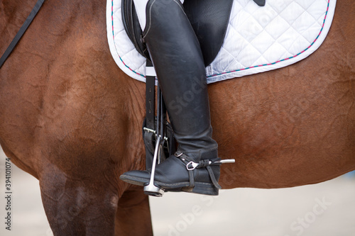 Croup of a red horse with a white saddle and a rider’s foot in a boot with a spur inserted in a stirrup.