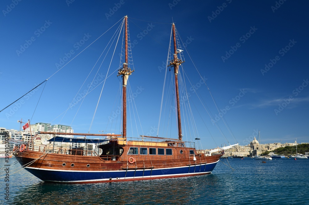 Obraz premium Cruise sailboat in the port of Tas Sliema, Valletta, Malta. A wooden sailboat waits in the harbor on a sunny day on the calm sea.