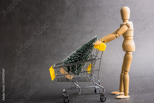 A wooden doll carries a Christmas treel in metal shopping cart. Concept of buying Christmas decorations, moneys, miniature. Stay on gray background. photo