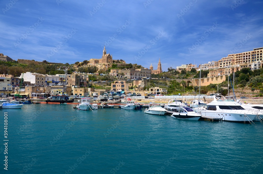 Panorama of the harbor on the island of Gozo, Malta. Boats in the harbor, church on the hill, sunny sky, Mediterranean atmosphere.