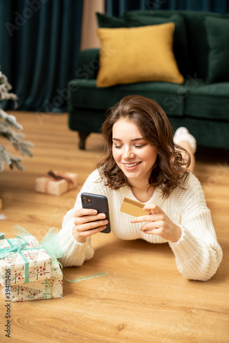 Smiling girl with smartphone and credit card near Christmas tree. Online shopping on new year. She buying presents