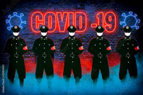 Silhouettes of Kovid cops with a mask in a colorful comic fog against the backdrop of a neon glowing inscription with icons and signs of the crown virus in blue and red hanging on a brick ruined wall