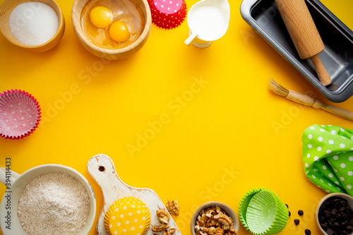 Baking frame with flour, sugar, milk, eggs, wallnuts, chocolate chips, rolling pin, baking mold. Yellow background with copy space.