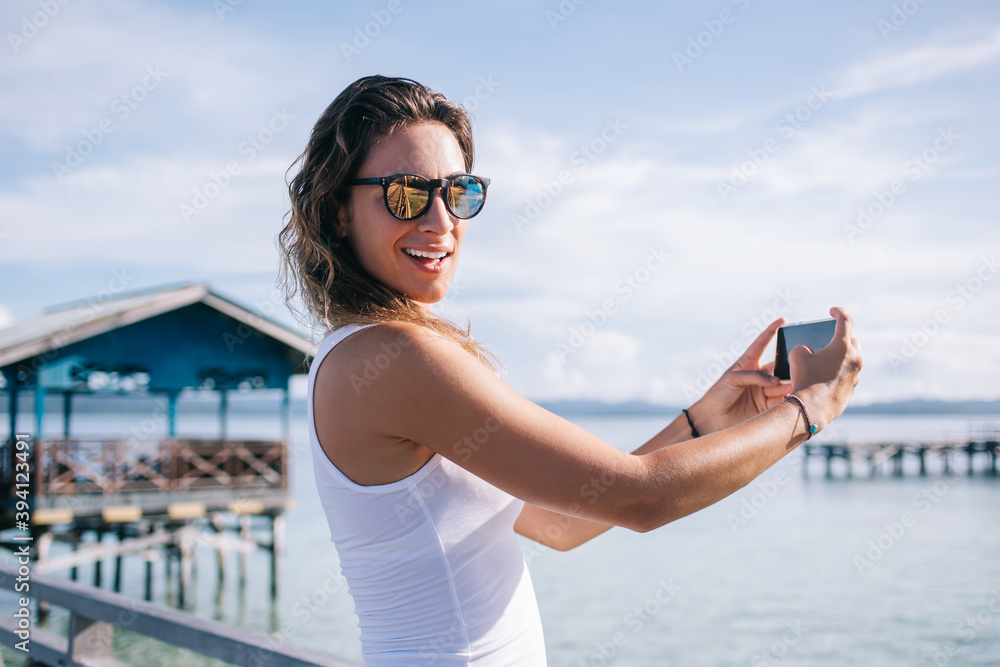 Half length portrait of happy female tourist in sunglasses smiling at camera while using cellular application for shooting live streams in social networks,millennial tourist enjoying vacations in Asia