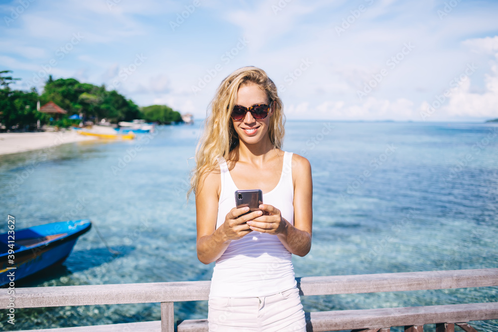 Cheerful woman in sunglasses enjoying smartphone messaging with social followers communicate about Maldives vacations, smiling female tourist typing text publication for sharing to travel web blog