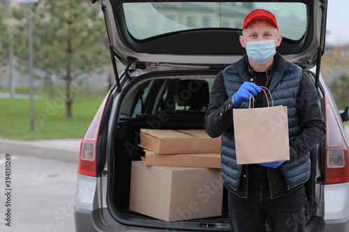 Delivery man in red cap, face medical mask and gloves hold paper bag near the car outdoor. Service coronavirus. Online shopping. mock up.