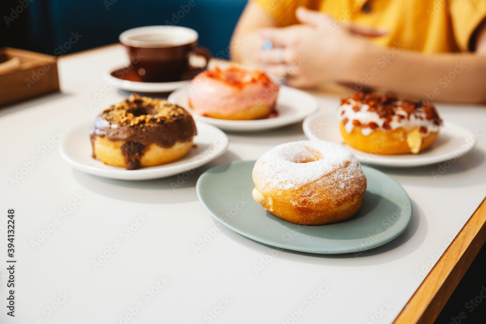 Donuts on the table include Strawberry pink, chocolate, sugar glazed and bacon, cheese donut on the table with the woman wearing yellow shirt in background in modern cafe. Enjoyment female lifestyle. Donuts on the table include Strawberry pink, chocolate, sugar glazed and bacon, cheese donut on the table with the woman wearing yellow shirt in background in modern cafe. Enjoyment female lifestyle.