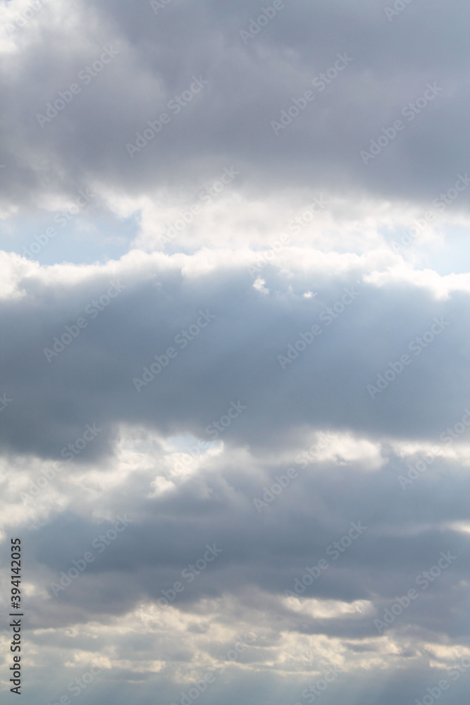 The blue sky is covered with dark clouds, close-up, bottom view. The rays of the sun shine through the gray clouds. Large clouds are arranged in three horizontal lines. Autumn day.