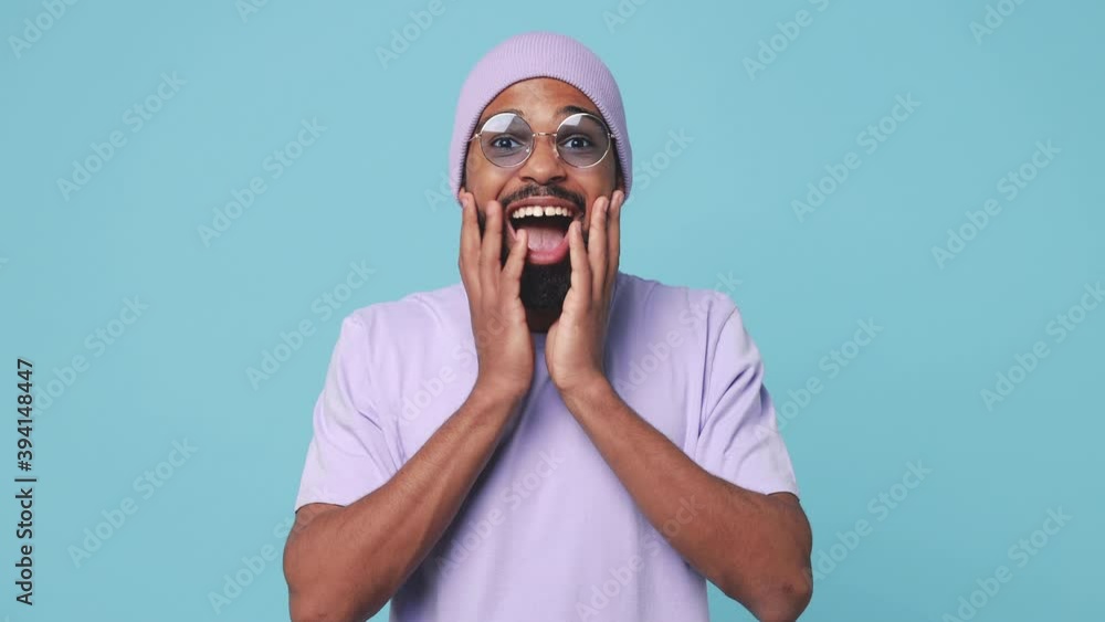 Shocked surprised young african american man 20s in violet t-shirt hat glasses isolated on blue turquoise background studio. People lifestyle concept. Looking camera say wow covering mouth with hands Shocked surprised young african american man 20s in violet t-shirt hat glasses isolated on blue turquoise background studio. People lifestyle concept. Looking camera say wow covering mouth with hands