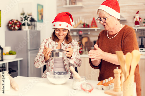 Grandchild playing with flour in christmas day. Happy cheerful joyfull teenage girl helping senior woman preparing sweet cookies to celebrate winter holidays wearing santa hat.