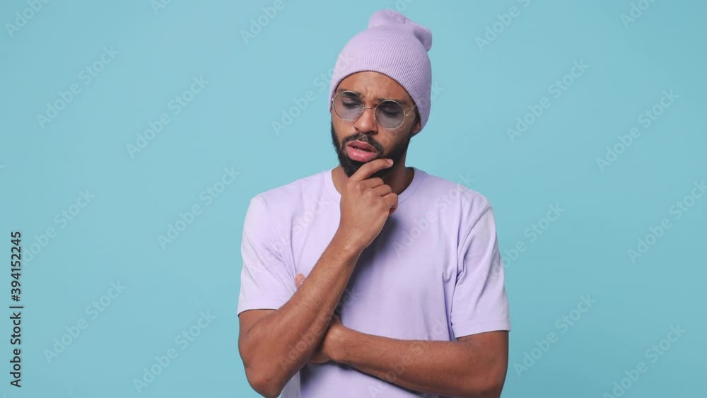 Confused puzzled young african american man 20s wearing violet t-shirt hat glasses isolated on blue turquoise background in studio. People lifestyle concept. Put hand prop up on chin spreading hands Confused puzzled young african american man 20s wearing violet t-shirt hat glasses isolated on blue turquoise background in studio. People lifestyle concept. Put hand prop up on chin spreading hands