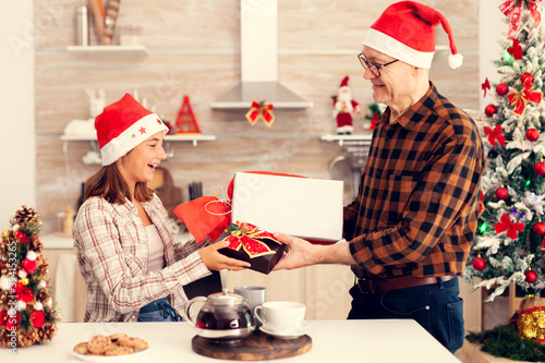 Happy grandchild exchanging presents with grandfather celebrating christmas. Senior man wearing santa hat surprising granddaughter with winter holiday gifts in home kitchen with xmas tree in the