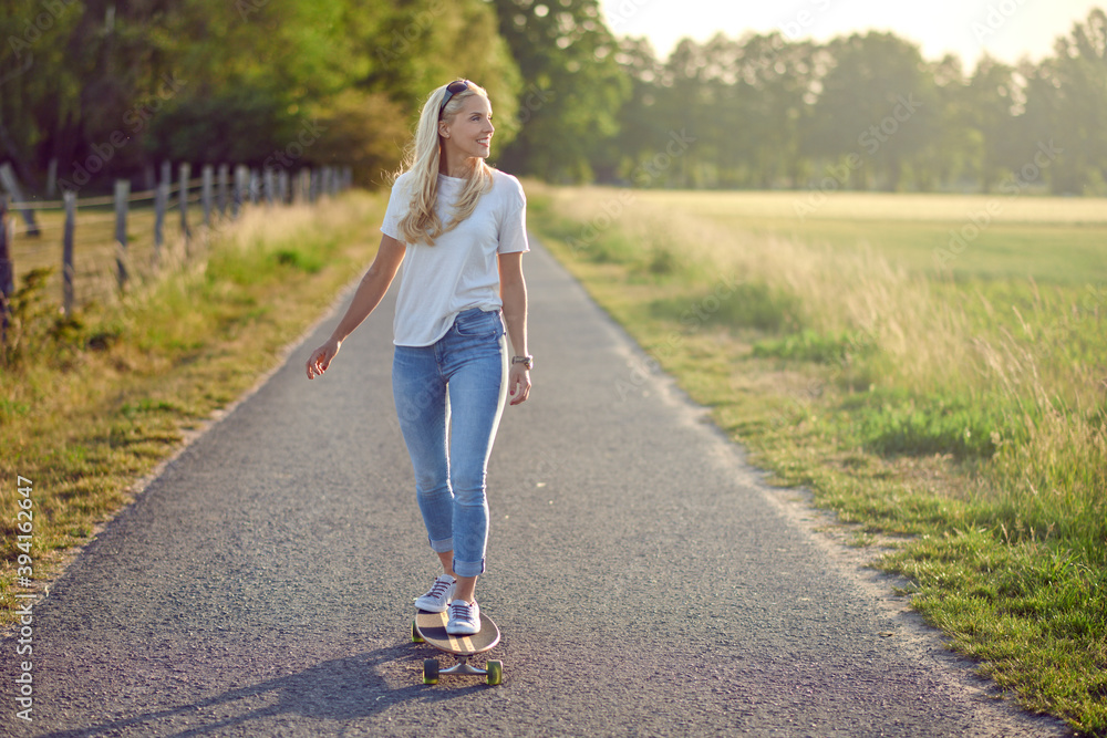 Fit active middle-aged woman playing on her skateboard approaching the camera along a narrow rural road with a happy smile backlit by the evening sun