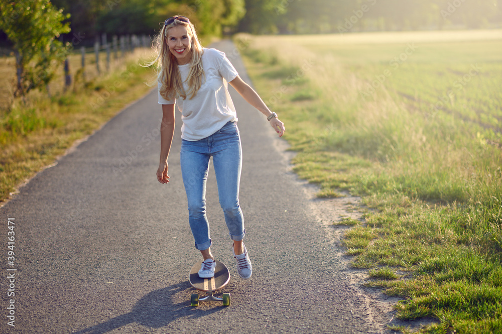 Fit active middle-aged woman playing on her skateboard approaching the camera along a narrow rural road with a happy smile backlit by the evening sun Fit active middle-aged woman playing on her skateboard approaching the camera along a narrow rural road with a happy smile backlit by the evening sun