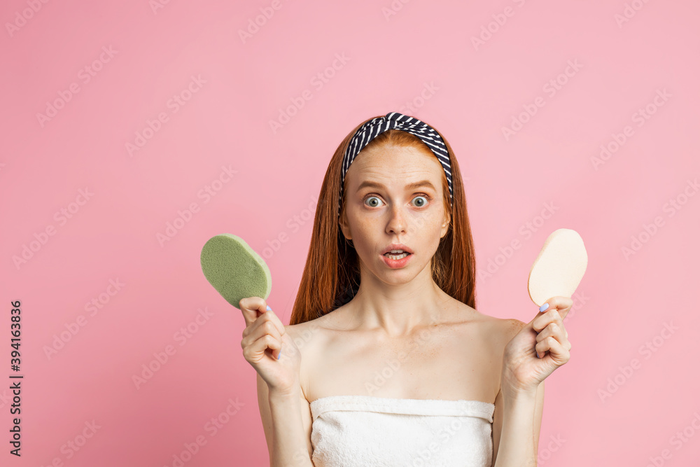 Closeup of stunned young ginger lady, wearing striped headband, staring at camera with open mouth, wrapped in white towel, holding two sponges, posing over pink studio wall. Closeup of stunned young ginger lady, wearing striped headband, staring at camera with open mouth, wrapped in white towel, holding two sponges, posing over pink studio wall.