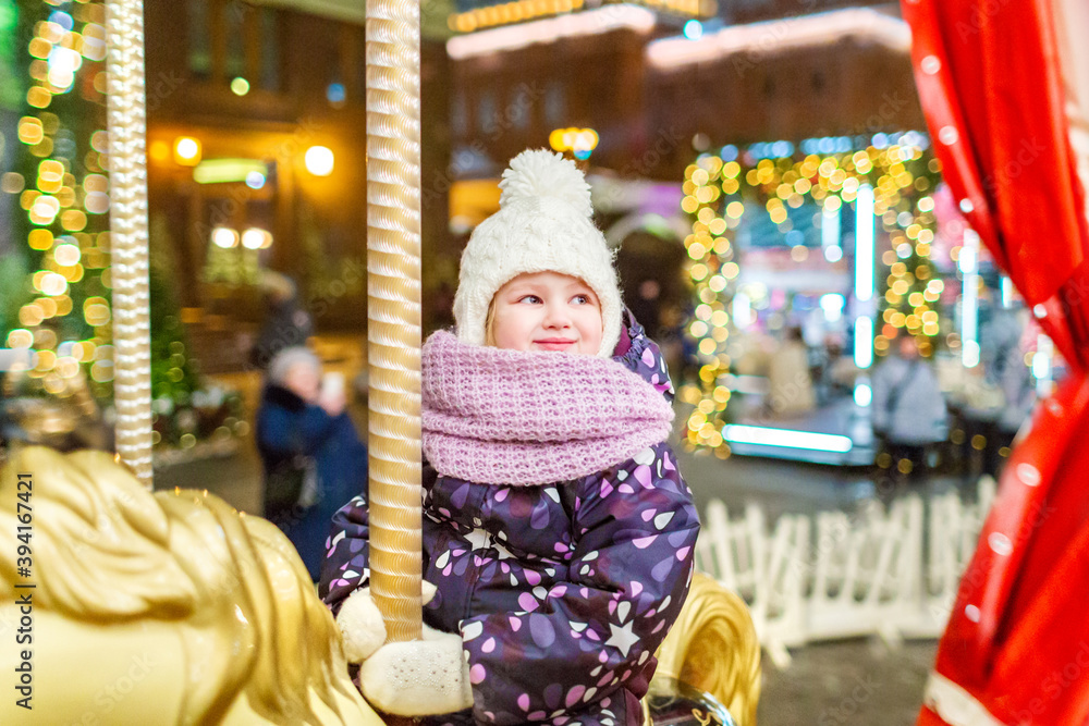 A little girl in warm clothes rides on a Christmas carousel with lights. Christmas market, night time