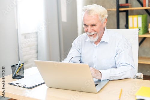 Concentrated grey-haired senior office employee using laptop sitting in the contemporary office space