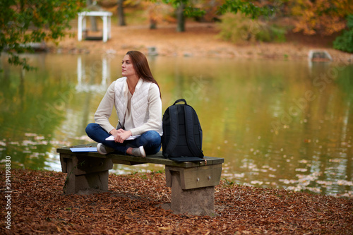 Beautiful young college student sits alone on campus in fall leaves near small pond - writing in notebook looking off frame and thinking