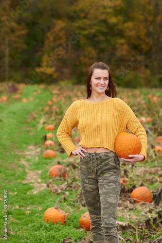 Adorable young woman in yellow crop sweater and camouflage pants stands in a pumpkin patch on farm - autumn holidays thanksgiving or Halloween themed - holding pumpkin against hip
