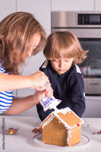 Boy child with grandmother or mother decorating a Christmas gingerbread house during the holidays using icing and white frosting as well as colorful sprinkles, candy cane, and gummy candy in kitchen