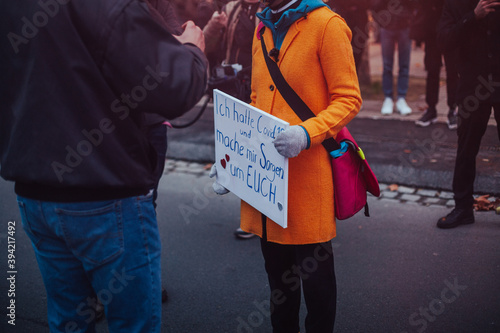 Woman in bright orange coat holding a sign is speaking with a demonstrator on a conspiracy theory demonastration. Her sign says 