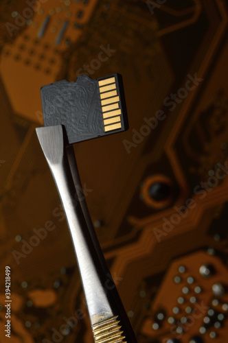 Close-up of a Micro SD memory card against the background of a microcircuit, clamped with tweezers.