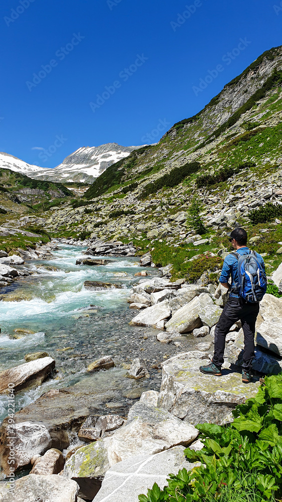 A man with a big backpack enjoying a cascading waterfall in the Alps in Ankogel group in Austria. The rushing torrent is coming from a glacier water. High mountains around, overgrown with small bushes