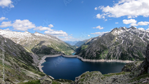 A panoramic view on an artificial  dam lake stretching over a vast territory around Alps in Austria. Lake is shining with navy blue color. In the back there are a few glaciers. Controlling the nature