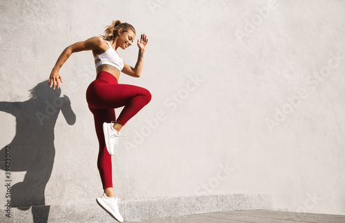 Fitness woman doing cardio interval training outdoors. Caucasian female in sportswear exercising outdoors in morning, jumping against concrete wall