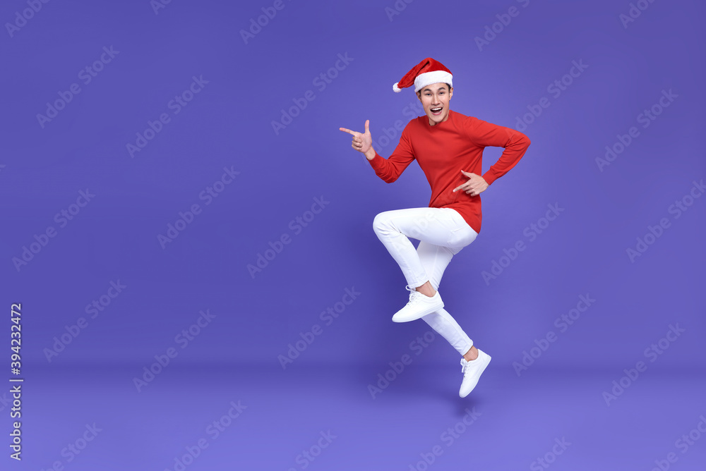 Young asian man in red casual attire wearing Santa hat jumping with pointing finger aside front a copy space and smile face on purple background.Happy new year concept.