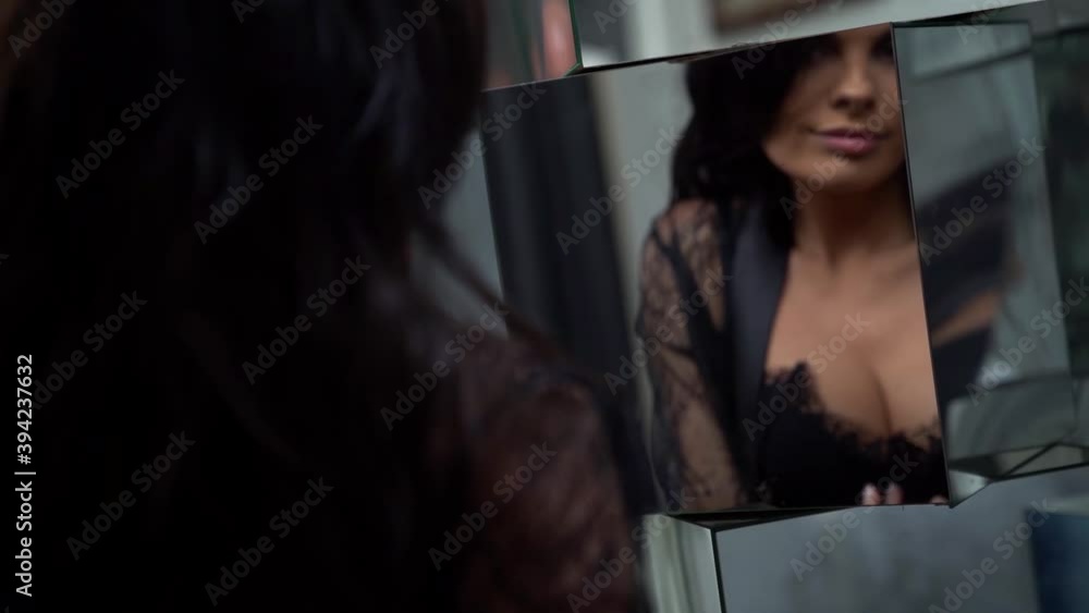 A brunette beauty in a black lace bodysuit that emphasizes her lush Breasts and seductively poses in a room in front of a mirror looking at her reflection. A brunette beauty in a black lace bodysuit that emphasizes her lush Breasts and seductively poses in a room in front of a mirror looking at her reflection.