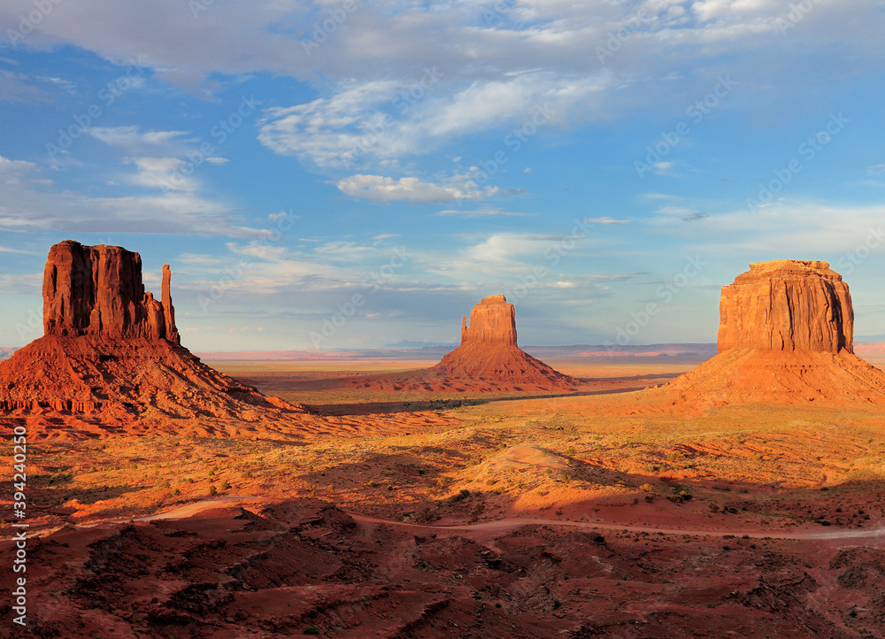 View To The East Mitten Butte, Merrick Butte And West Mitten Butte In The Monument Valley Arizona In The Late Afternoon Sun On A Sunny Summer Day With A Clear Blue Sky And A Few Clouds