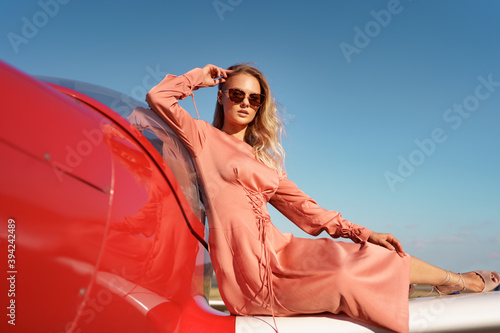 Pretty blond woman with wavy hair wearing pink silk classy dress posing near a private plane © diignat