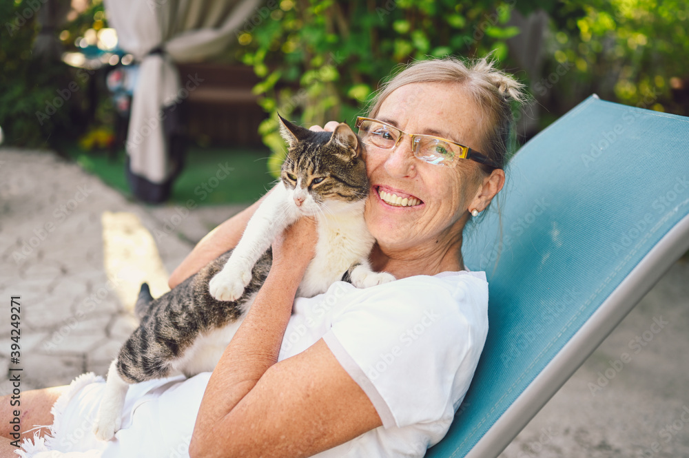 Obraz premium Happy smiling senior elderly woman in glasses relaxing in summer garden outdoors hugging domestic tabby cat. Retired old people and animals pets concept