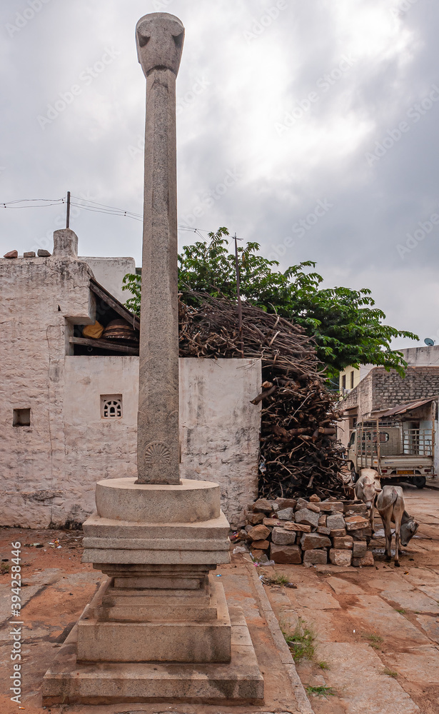 Kaddirampura,, Karnataka, India - November 4, 2013: Lone gray stone pillar in center of town as remnant of ancient Shiva sanctuary and first adorations of the Hindu gods. Gray sky and some green folia
