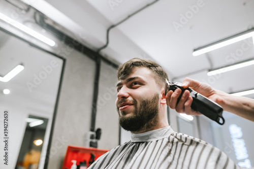 Funny bearded man in peignoir sitting in hairdresser chair and haircut in barber with a smile on his face, looking away, close portrait. Funny adult client cuts beard and head in hairdresser.