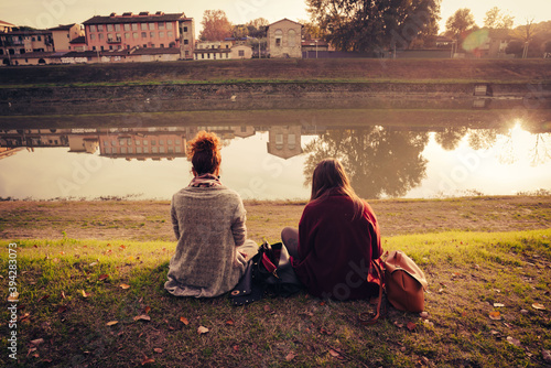 two young female friends sitting on the bank of a river while using their smartphones