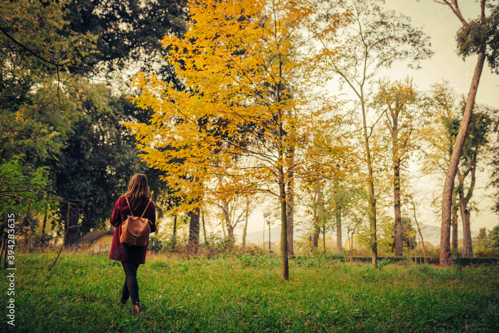 Obraz premium young woman with red coat and leather backpack while walking in the middle of a city park with autumn colors