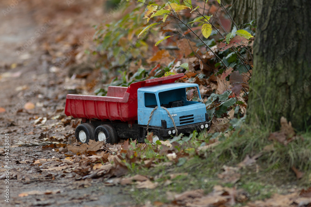 Red and blue toy industrial truck vehicle left behind on the side of a pathway filled with autumn colored leaves